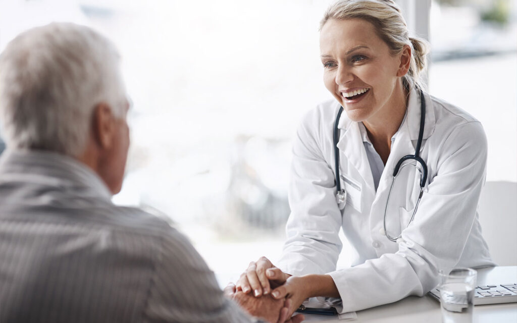 Doctor holding a patient’s hands during a caring visit, representing relationship-based primary care and how we return to healing